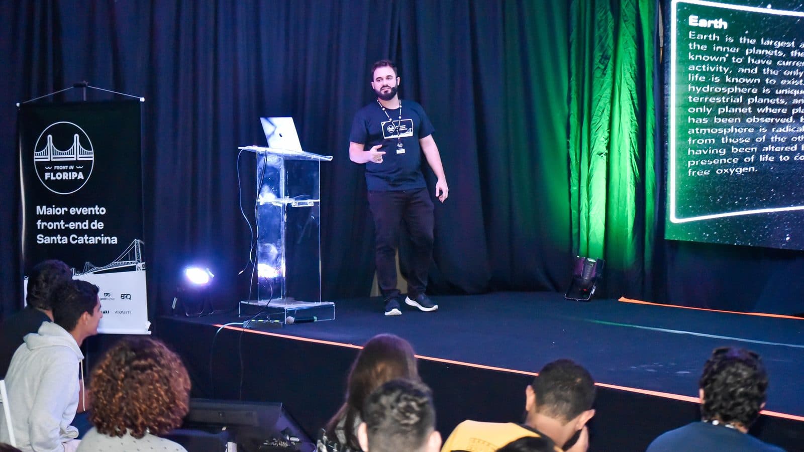 Eduardo Sotero on stage at Front in Floripa 2019, mid-sentence beside a lectern, with the conference banner reading "Maior evento front-end de Santa Catarina" to his left and a slide from the Solar System demo projected to his right.